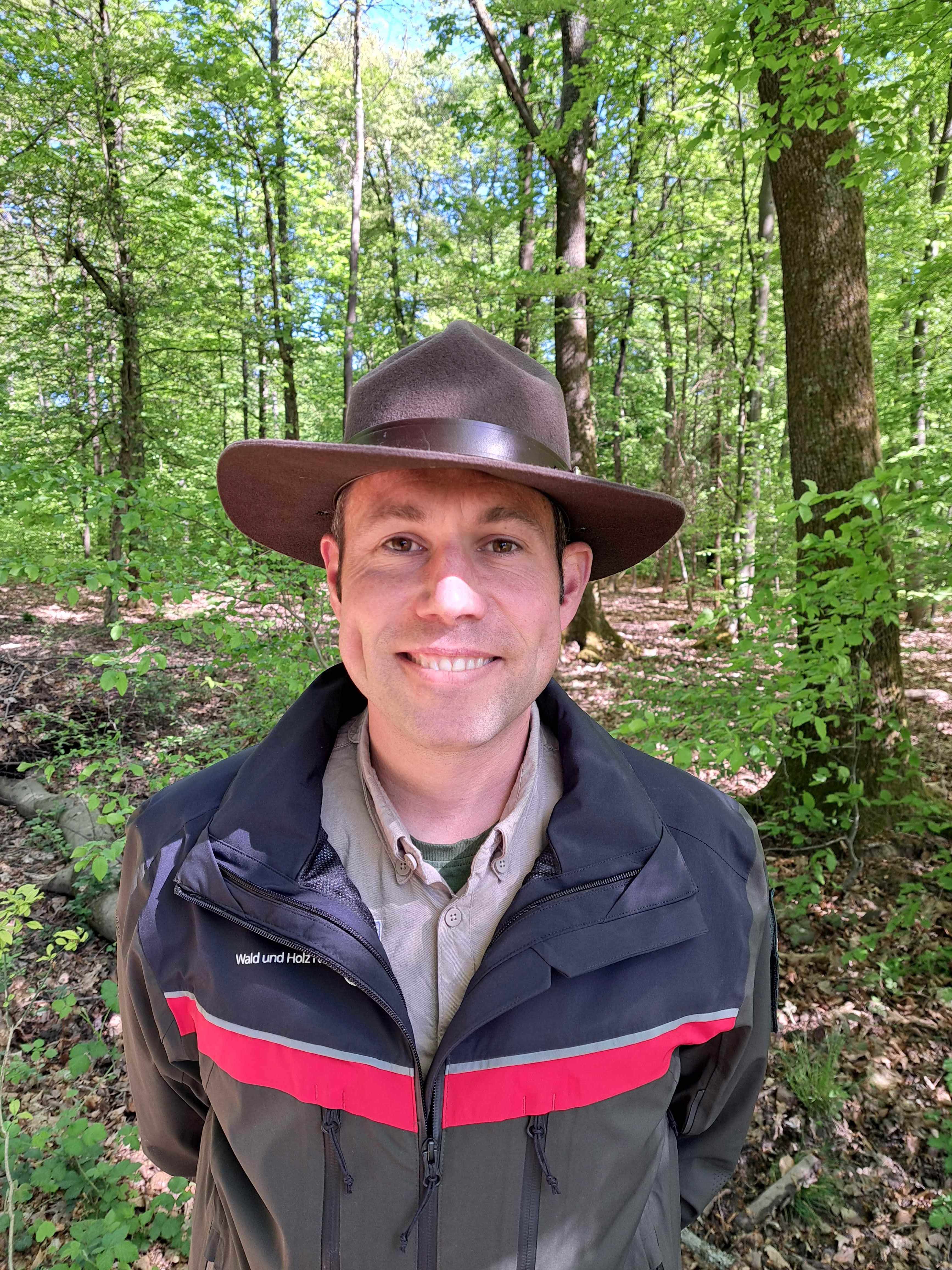 Mann in grün-schwarz-roter Dienstkleidung und Filzhut steht im Wald und schaut freundlich in die Kamera. 