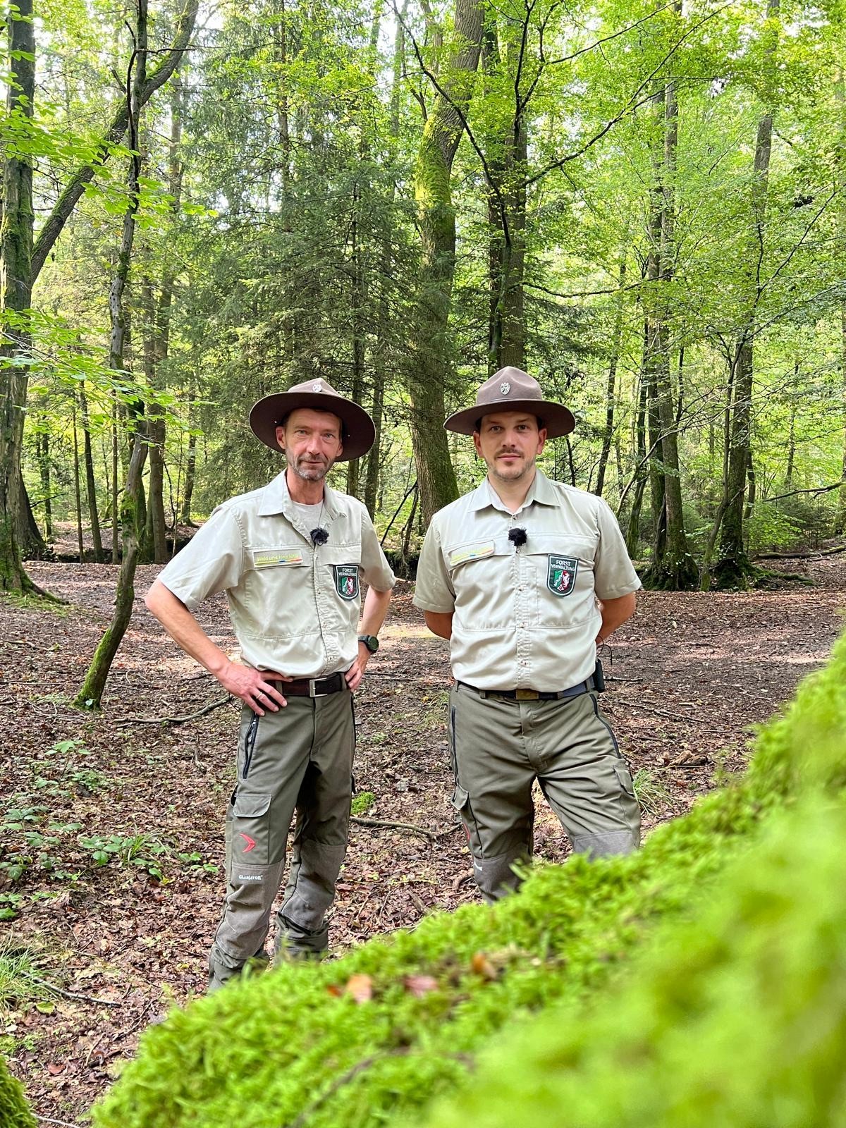 Zwei Männer stehen im Wald und schauen freundlich in die Kamera. Sie tragen forstliche Dienstkleidung und einen Hut.