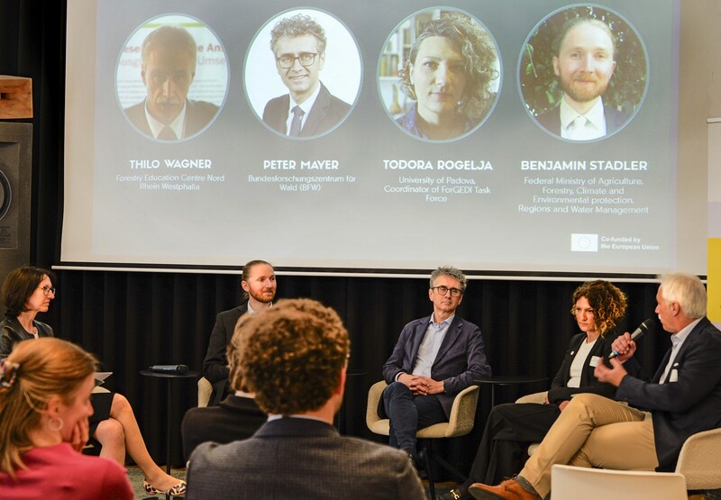 Podiumsdiskussion mit mehreren Expertinnen und Experten vor einer Präsentation mit Porträts und Namen der Teilnehmenden.